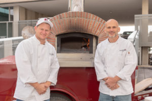 Two pizza makers in chef coats standing in front of a mobile wood-fire pizza oven
