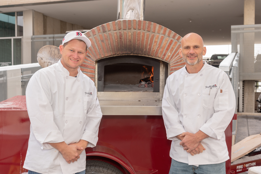 Two pizza makers in chef coats standing in front of a mobile wood-fire pizza oven